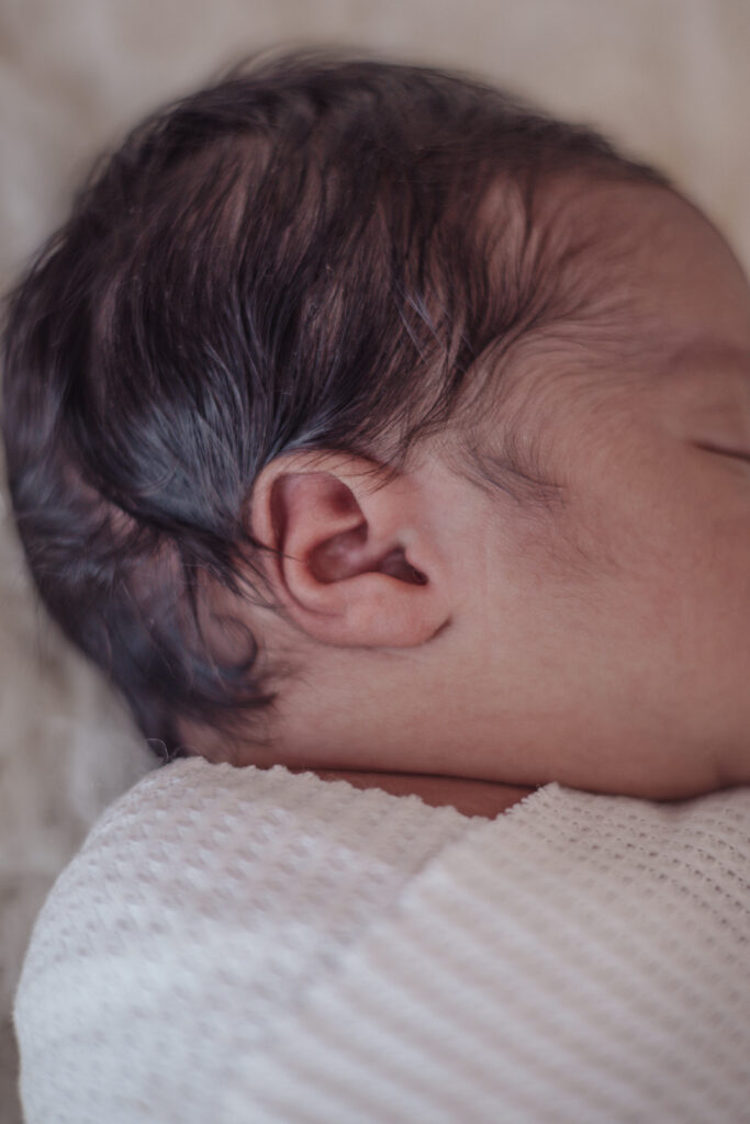Gros plan de la tête d’un nouveau‑né enveloppé dans une couverture blanche, séance lifestyle en lumière naturelle.