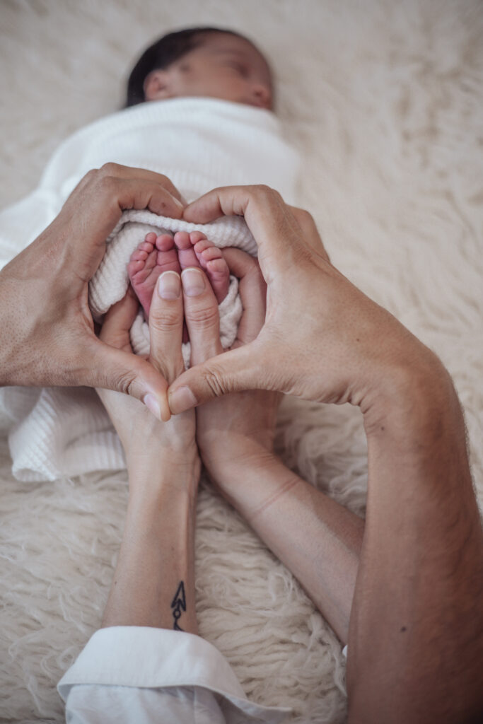 Pieds de nouveau‑né entourés des mains de ses parents, moment tendre capturé en séance lifestyle à domicile.