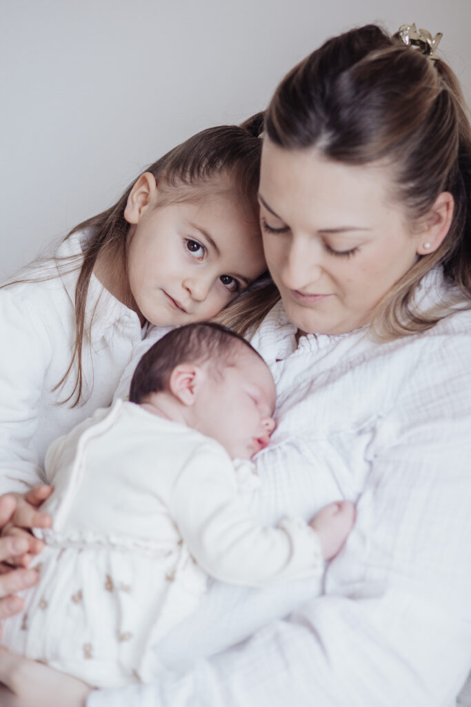 Jeune maman tenant son nouveau‑né avec sa fille à ses côtés, moment tendre en lumière naturelle.