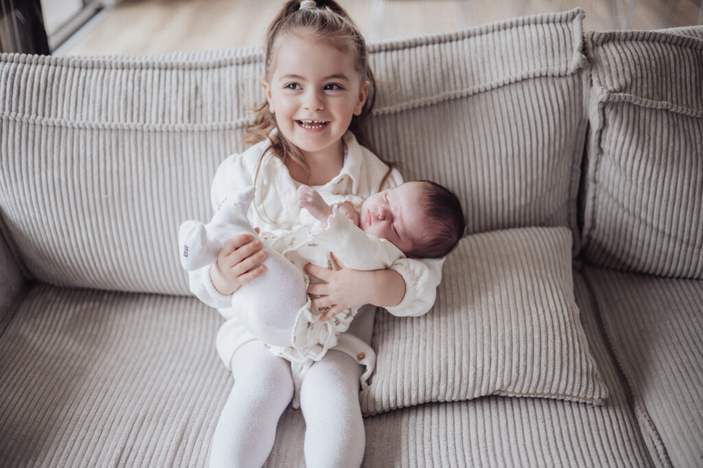 Grande sœur embrassant son nouveau‑né dans les bras d’un parent, moment tendre en lumière naturelle.