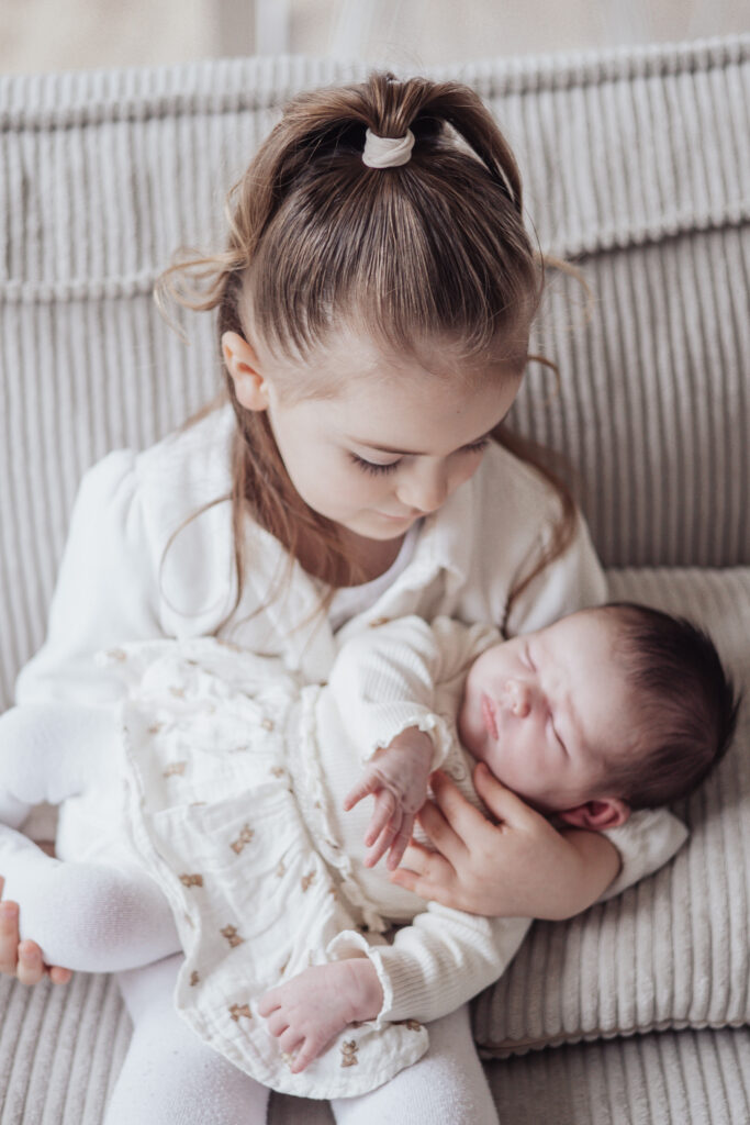 Grande sœur berçant son nouveau‑né sur le canapé, scène douce et intime en lumière naturelle.