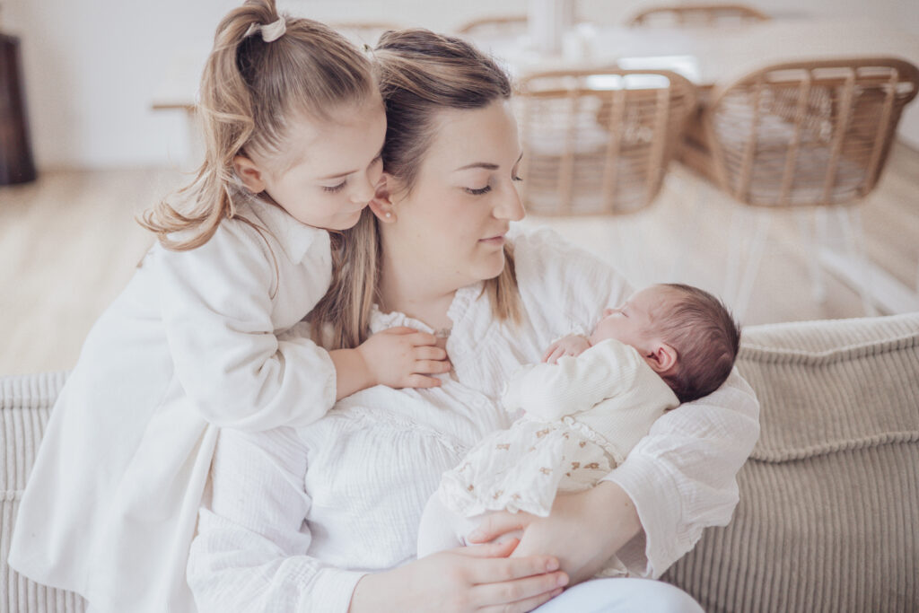 Grande sœur tenant son nouveau‑né sur le canapé, scène lumineuse et pleine de complicité.