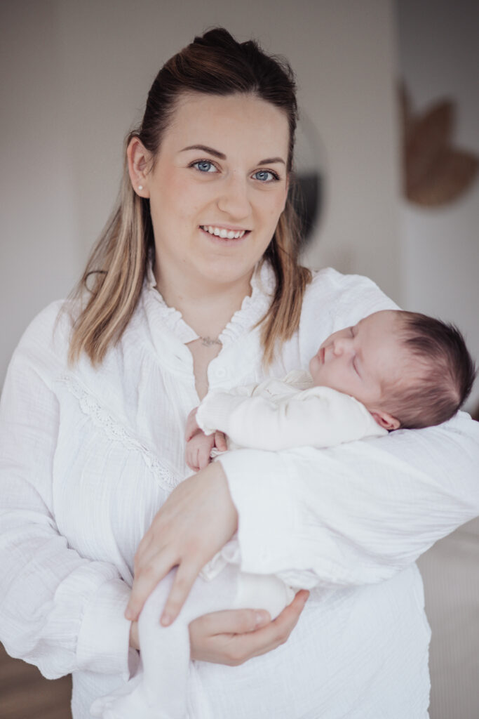 Jeune maman debout tenant son nouveau‑né endormi, scène lumineuse et naturelle.