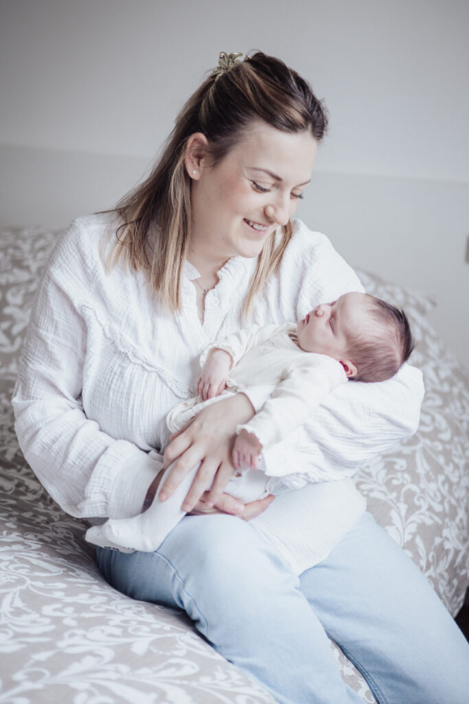 Jeune maman assise sur le lit tenant son nouveau‑né endormi, moment doux en lumière naturelle.