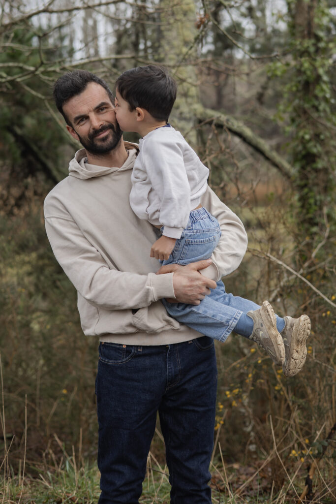 Papa tenant son enfant et recevant un bisou sur la joue, moment tendre en extérieur dans la forêt.