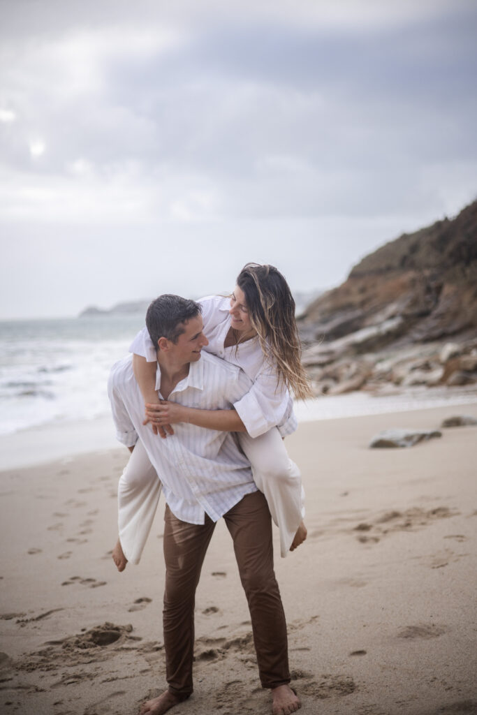 Couple jouant sur la plage, la femme sur le dos de son partenaire, moment complice en lumière naturelle.
