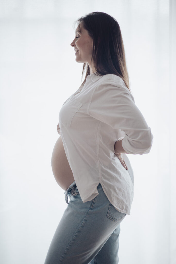 Portrait de future maman en intérieur, chemise blanche ouverte sur le ventre, photographie grossesse lifestyle.