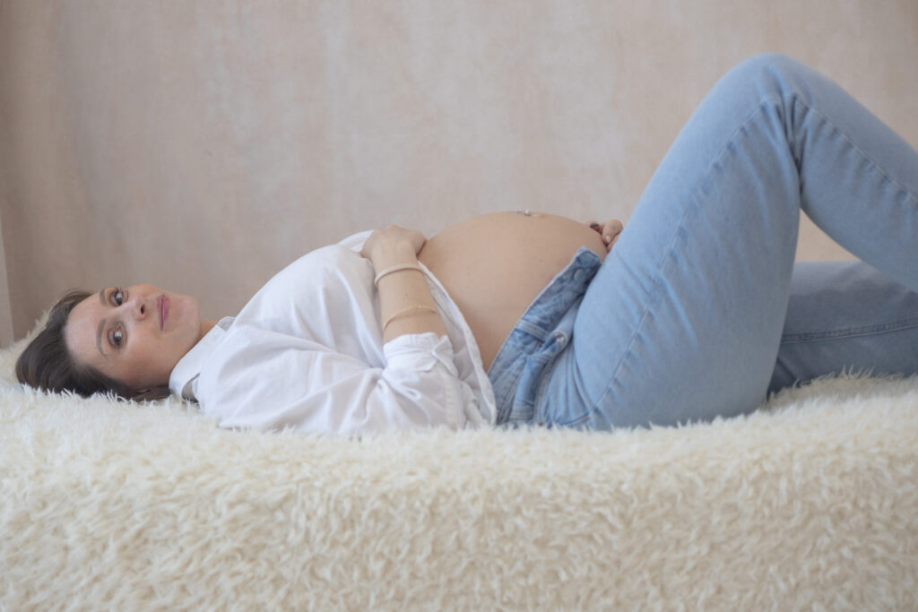 Femme enceinte allongée sur une surface douce, mains posées sur son ventre, séance grossesse en lumière naturelle.