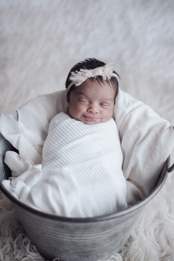 Nouvelle‑née endormie dans un seau, enveloppée dans une couverture blanche, séance photo nouveau‑né lifestyle.