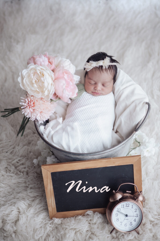 Nouvelle‑née posée dans un seau avec fleurs et ardoise prénom, séance nouveau‑né artistique en lumière naturelle.