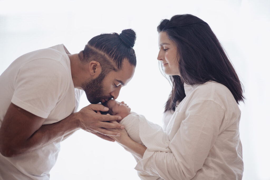Parents tenant leur nouveau‑né en lumière naturelle, moment tendre avec un bisou sur la tête. 